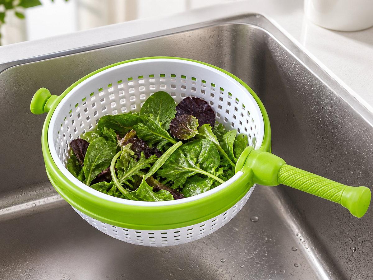 Green and white handheld salad spinner with rinsed leafy greens over a kitchen sink