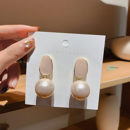A hand holds a white display card featuring a pair of drop earrings with oval blush pink tops and large round pearl-like beads in a soft champagne tone. The card is branded 'poparozzi'.