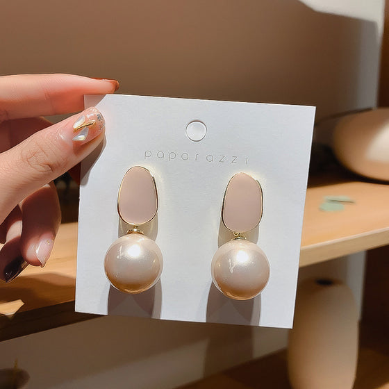 A hand holds a white display card featuring a pair of drop earrings with oval blush pink tops and large round pearl-like beads in a soft champagne tone. The card is branded 'poparozzi'.