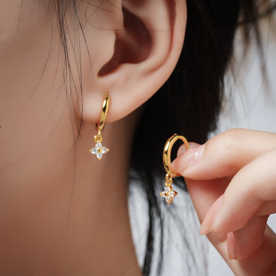 Gold-tone huggie hoop earrings with dangling four-leaf clover charms, worn on the ear