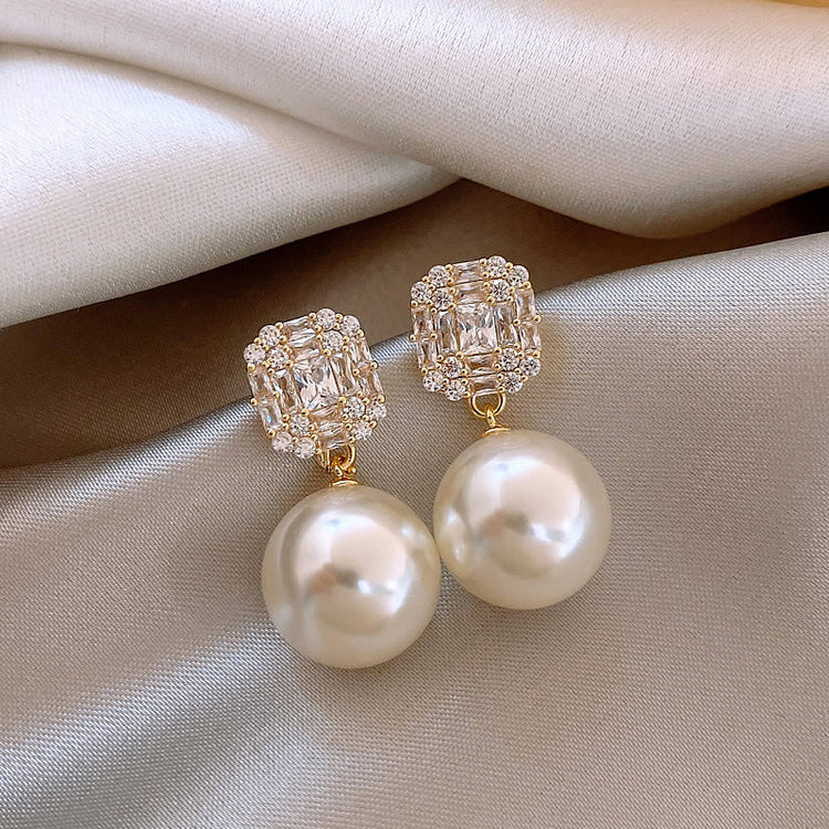 Pair of gold-tone copper stud earrings with a square zircon cluster and faux pearl drop, laid on fabric