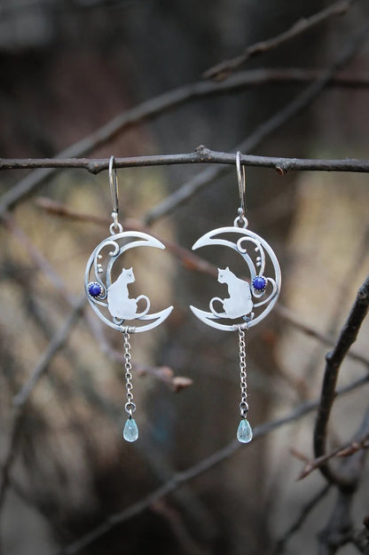 Silver-tone crescent moon and cat charm dangle earrings with blue bead drops, laid on dark stone with a black starfish accent