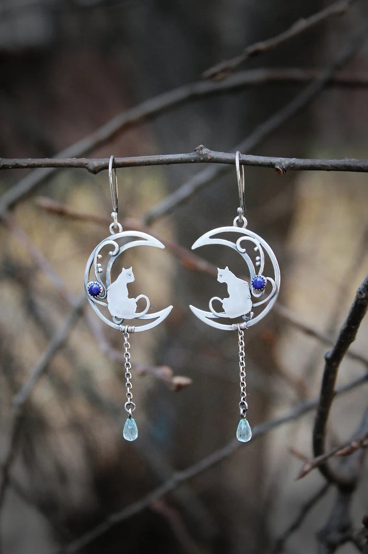 Silver-tone crescent moon and cat charm dangle earrings with blue bead drops, laid on dark stone with a black starfish accent
