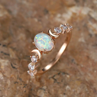 Rose gold-tone ring with an oval opal-style center stone and crescent moon accents, angled view on a stone surface